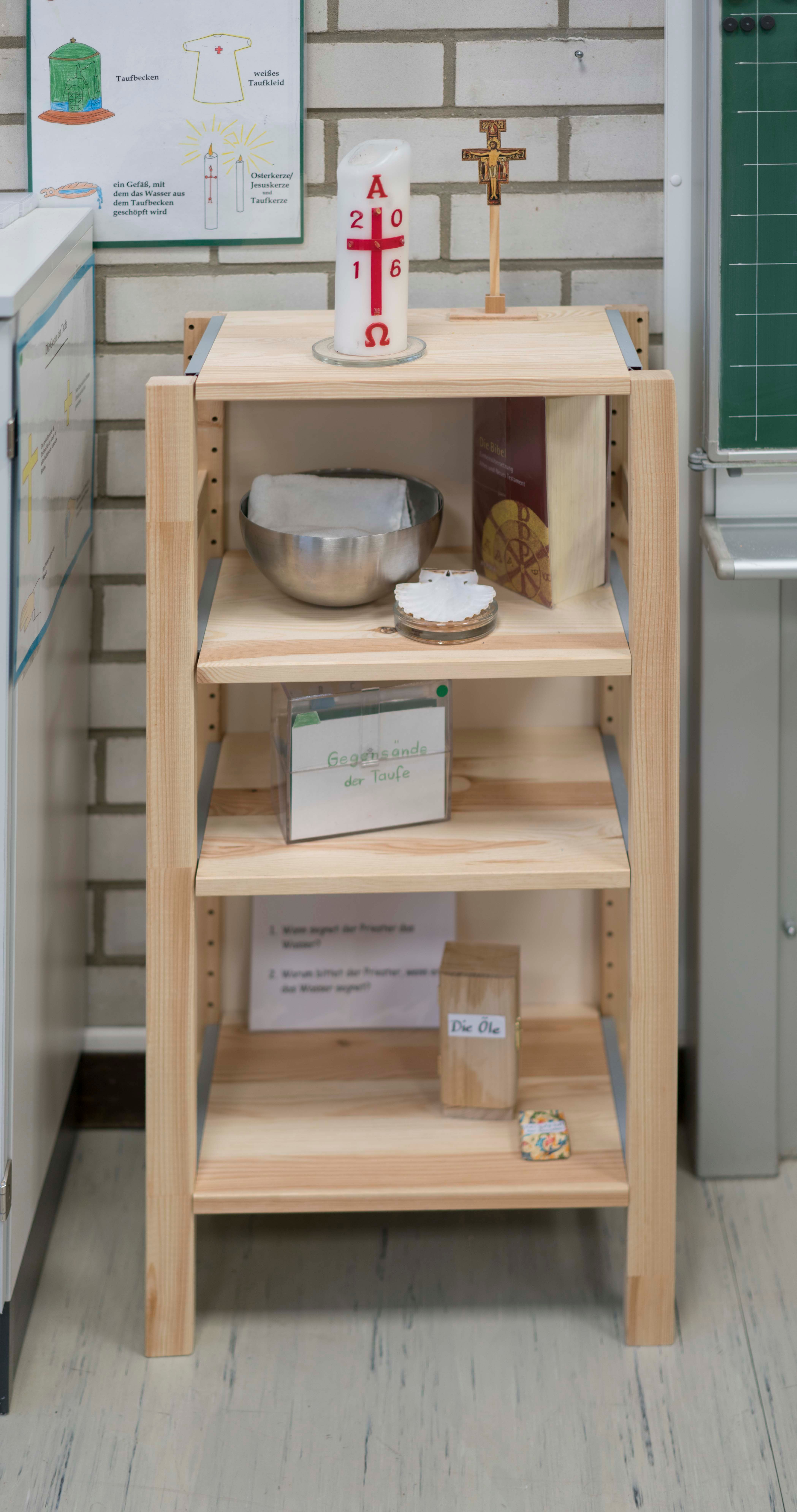 Die Taufecke besteht aus einem offenen Regal mit Osterkerze, Kreuz, Taufbecken, Bibel und weiteren Materialien zur Taufe.