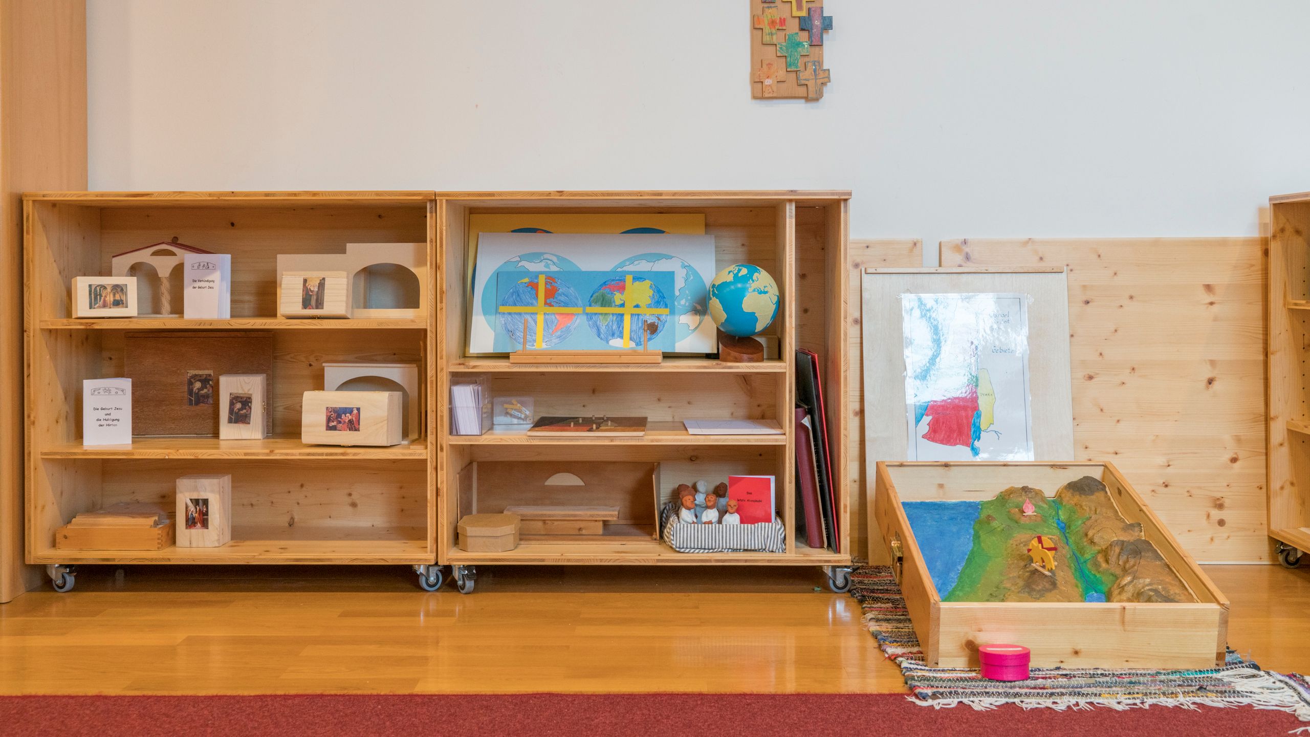 Offene, rollbare Regale mit kleinen Büchern mit Bibeltexten, dazu Figuren und kleinen Häusern, das Relief Israels und Landkarten