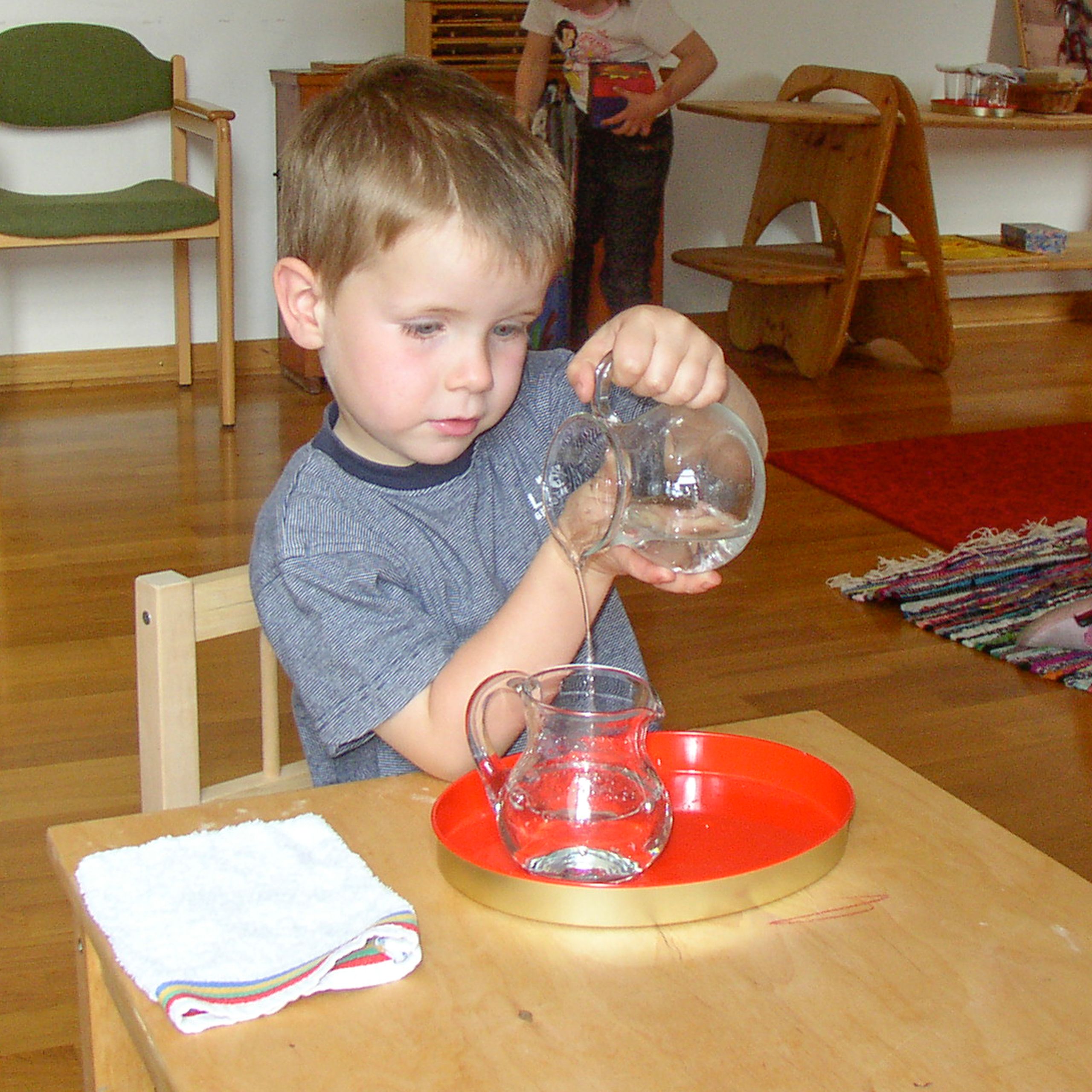 Kleiner Junge gießt konzentriert Wasser aus einem Kännchen in ein anderes Kännchen