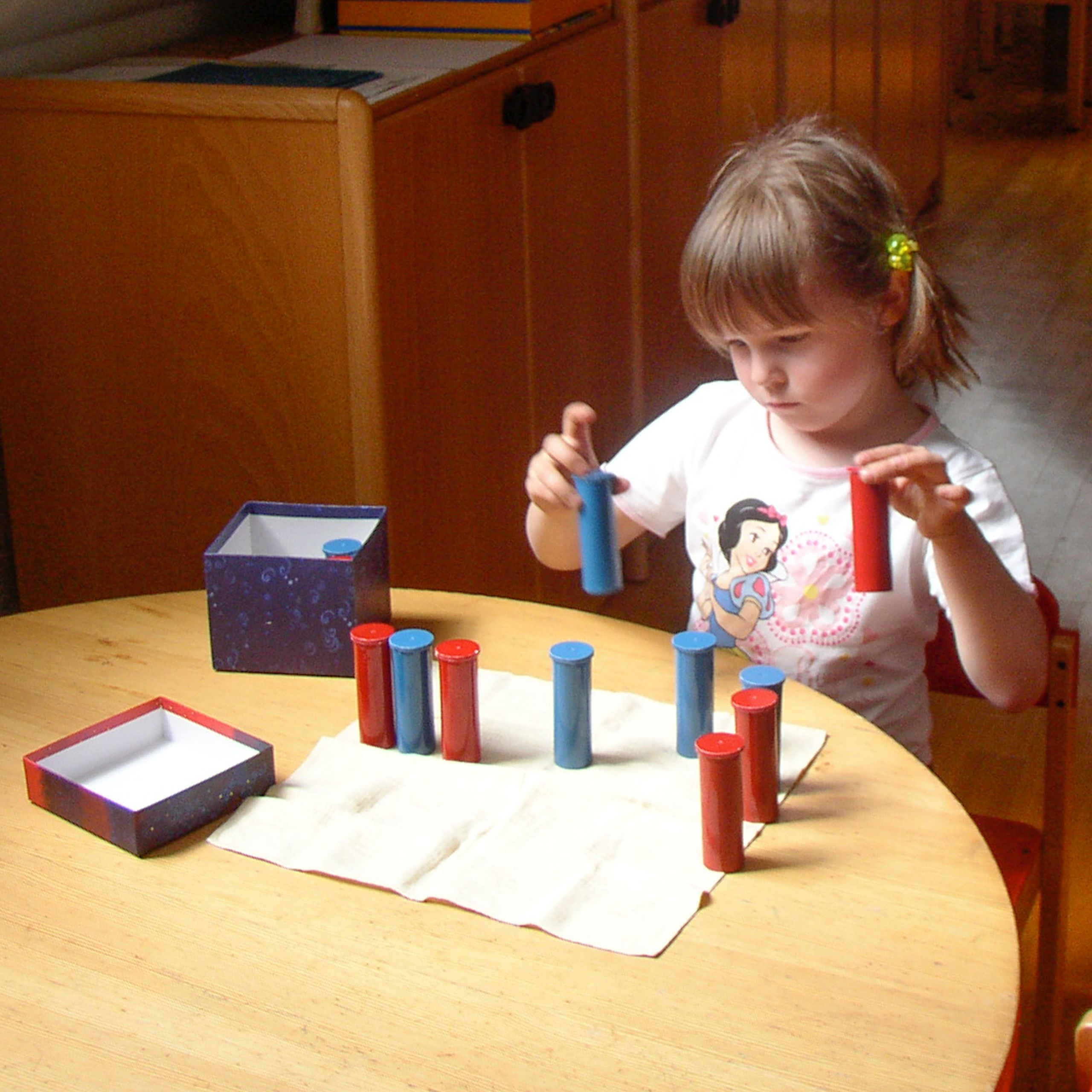 Ein kleines Mädchen schüttelt blaue und rote Geräuschdosen und vergleicht das Geräusch.